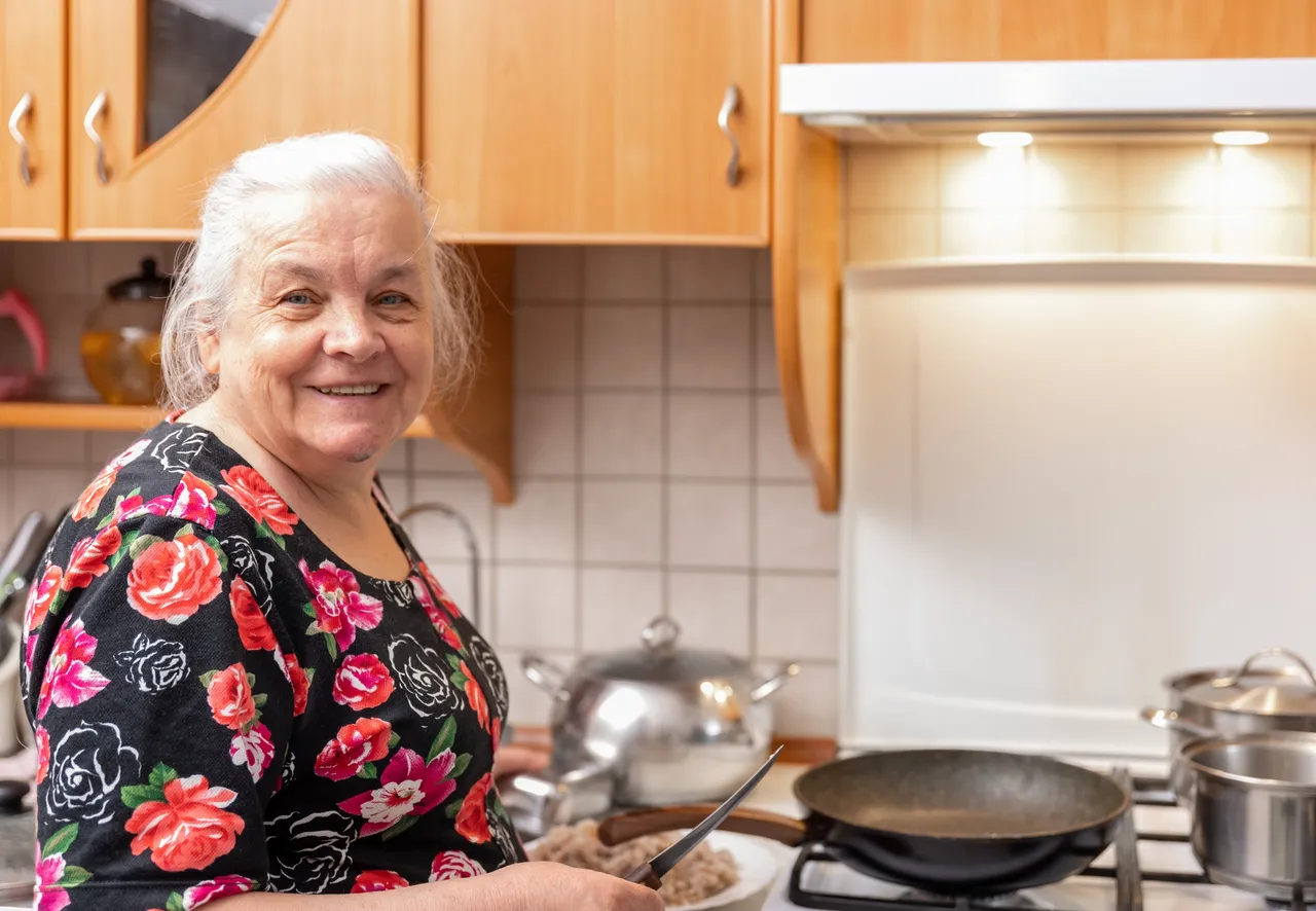An old woman with a smile on her face is standing in the kitchen near the stove cooking dinner.